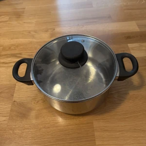 Kastrull med lock - Bilderna visar en kastrull i rostfritt stål med svarta handtag och ett tillhörande lock. Kastrullen ser rymlig ut och passar perfekt för att laga mat till flera personer. Perfekt för dig som gillar att experimentera i köket eller vill ha en pålitlig kastrull till vardagsmatlagningen.