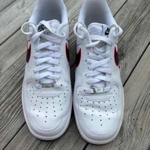 Nike Air Force 1 vita sneakers med röd och marinblå detalj - Säljer ett par knappt använda klassiska Nike Air Force 1 sneakers i vitt skinn med röd Swoosh och marinblå detaljer. Skorna har perforerad tå, vit snörning och mörkblå sula. På hälen står det '97 '98' och '99 '00' broderat i rött. 