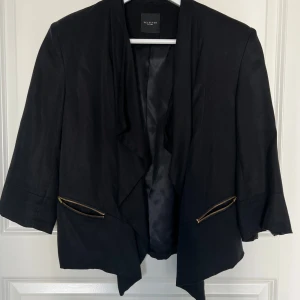 Svart kavaj från Selected Femme - Stilren svart kavaj från Selected Femme med öppen front och guldfärgade dragkedjor vid fickorna (obs mkt små fickor!). Kavajen har trekvartsärm och en mjuk, något lös passform. Perfekt för en trendig och uppklädd look.
