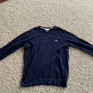 Lacoste tröja  - Säljer en klassisk mörkblå tröja från Lacoste med rund halsringning och det ikoniska krokodilmärket broderat på bröstet. Tröjan har långa ärmar och ribbade muddar vid ärmslut och nederkant. Perfekt för en stilren look.