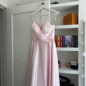 Ljusrosa långklänning med snörning i ryggen - Säljer en ljusrosa långklänning med smala axelband och v-ringad framtill. Klänningen har en vacker snörning i ryggen och är i satin material. Perfekt för bal eller festliga tillfällen. Klänningen är aldrig använd utan bara provad hemma. 