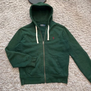 Grön hoodie från Polo Ralph Lauren - Mörkgrön hoodie med dragkedja från Polo Ralph Lauren. Tröjan har huva med vita snören, två fickor framtill och en liten rosa broderad logga på bröstet. Perfekt för en avslappnad stil. Väldigt bra skick förutom skadorna vid händerna. Ungefär samma sak på andra handen. Fråga gärna om närmare bilder på etiketten och loggan. Skriv om du undrar över något annat!