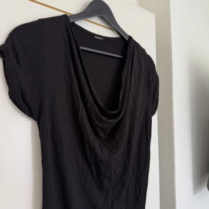 Svart klänning med draperad hals - En svart klänning med korta ärmar och snygg draperad halsringning. Modellen är enkel och stilren, perfekt för dig som gillar minimalistisk stil. Klänningen har en rak passform och är tillverkad i ett mjukt material.
