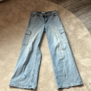 Ljusblå cargobyxor från Lindex - Säljer ett par ljusblå cargobyxor från Lindex med breda ben och flera fickor på sidorna. Byxorna har normal passform och är tillverkade i jeansmaterial. Perfekta för en avslappnad stil. Har defekter längst ner på benen som är färg. Hör av er vid frågor❤️