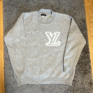 Grå stickad tröja från Louis Vuitton - Snygg grå stickad tröja från Louis Vuitton med stort vitt LV-logo på bröstet. Tröjan har rund halsringning och ribbade muddar vid ärmslut och nederkant. Perfekt för dig som vill ha en exklusiv och stilren look.