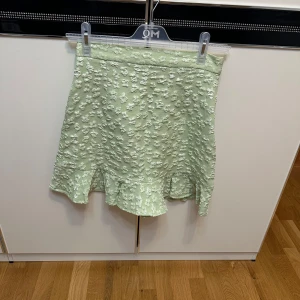 Ljusgrön kjol med volang - Superfin ljusgrön kjol med diskret blommönster och volang nertill. Kjolen har en lätt glansig yta och är perfekt för dig som vill ha något unikt och fräscht i garderoben. Stängs med dragkedja i sidan.
