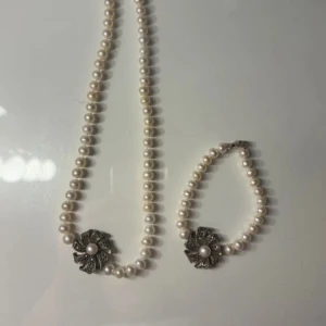 Pärlset halsband och armband med blomdetalj - Säljer ett matchande set med halsband och armband av vita pärlor. Båda smyckena har en dekorativ blomformad detalj i silverfärg med en pärla i mitten. Perfekt för att ge en elegant touch till din outfit.
