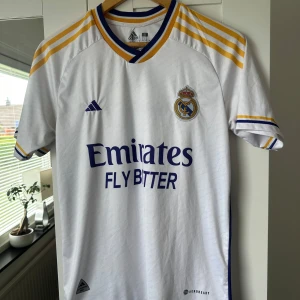 Real Madrid hemmatröja - Säljer en vit Real Madrid hemmatröja från Adidas med gula och blå detaljer. Tröjan har klubbmärke på bröstet, Adidas-logga och sponsortryck framtill. Klassisk design med korta ärmar och randiga axlar. 