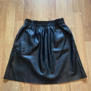 Svart kjol i skinnimitation från Vintage Dressing - Säljer en svart kjol i skinnimitation från Vintage Dressing. Kjolen har resår i midjan och är helsvart med en slät yta. Perfekt för dig som gillar en stilren look och vill ha något som sticker ut lite extra.