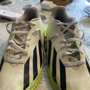 Adidas Crazyfast fotbollsskor i vitt och neongult - Säljer ett par Adidas Crazyfast fotbollsskor i vitt med neongula detaljer och svarta ränder. Skorna har snörning och platt sula, perfekt för snabba rörelser på planen. Klassisk Adidas-design med sportig känsla.