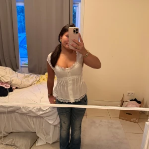 Vit spetsdetaljerad blus med knappar - Säljer en söt vit blus med spetsdetaljer och knappar framtill. Blusen har v-ringning, är ärmlös och har fina spetskanter längs halsringningen och nederkanten. Perfekt för dig som gillar romantisk och feminin stil.