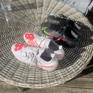 Fotbollsskor 41 Nike  - Säljer ett par vita New Balance fotbollsskor med röda detaljer och snörning. Skorna har en platt sula med dobbar och stora NB-loggor på sidorna. Perfekta för dig som vill sticka ut på planen. Samt ett pr svarta Nike mercurial