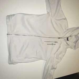 Vit hoodiejacka från Sail Racing - Säljer en vit hoodiejacka från Sail Racing med dragkedja framtill och en extra dragkedjeficka på bröstet. Jackan har huva och logga broderad på bröstet. Perfekt för dig som gillar stilren och sportig look. 176 storlek, men passar även på mig 180. Helt ny skicka förutom att färgen är inte lika vitt som om den hade varit nyköpt.