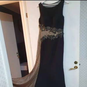 Svart långklänning med mesh och spetsdetaljer - Säljer en svart långklänning med meshpartier och dekorativa spetsdetaljer över byst och midja. Klänningen har ärmlös design och en lång, elegant siluett med transparent mesh längs sidan och ryggen. Perfekt för en speciell kväll!