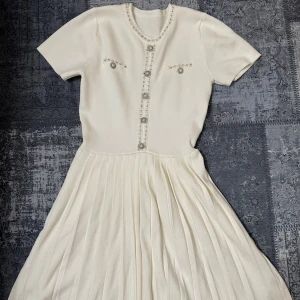Vit plisserad klänning med pärldetaljer - Säljer en elegant vit klänning med korta ärmar och plisserad kjol. Klänningen har fina pärldetaljer längs halsringningen och framtill, samt dekorativa knappar. Perfekt för sommarens tillfällen!