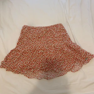 Rödblommig volangkjol - Supersöt kort kjol med rött blommönster och volangdetaljer. Kjolen har en luftig och flowy känsla, perfekt för varma dagar. Resår i midjan för extra komfort och snygg passform.