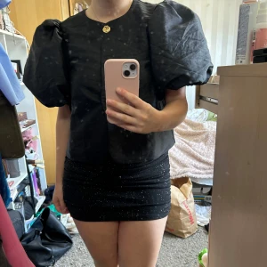 Svart glittrig minikjol - Säljer en svart glittrig minikjol. Kjolen är tight men väldigt stretchig och har en kort passform. Den kan justeras utan att det ser konstigt så den är både låg eller hög midjan beroende på hur man vill ha den. Endast använd en gång så i väldigt bra skick!