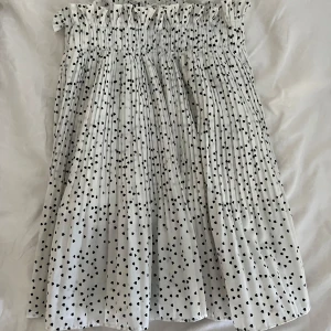 Vit tub topp med svarta hjärtan - Supersöt vit topp med svarta hjärtan och smock upptill. toppen har avskuren axel och ett luftigt fall, perfekt för varma dagar. Mjuk och lätt i materialet, med ett lekfullt mönster som sticker ut.