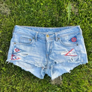 Ljusblå jeansshorts med patchar och slitningar - Säljer ett par ljusblå lågmidjade jeansshorts med slitningar och färgglada patches i rött, vitt och blått. 
