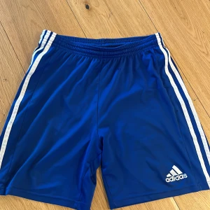 Blå Adidas shorts med vita ränder i storlek 176 - Snygga blå shorts från Adidas med klassiska vita ränder längs sidorna och Adidas-logga på ena benet. Perfekta för sport eller träning, med elastisk midja för skön passform. Materialet är lätt och andas.