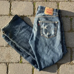 Levis 501 blå jeans med slitna detaljer - Klassiska Levis 501 jeans i blå denim med slitna detaljer och lite trasiga benslut. Byxorna har fem fickor, knappgylf och den ikoniska Levis-lappen bak i midjan. Perfekta för en avslappnad och tidlös stil.