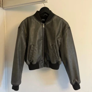 Grå khaki bomberjacka i skinnimitation från Zara - skinnjacka / bomberjacka i skinnimitation från Zara. Jackan har dragkedja framtill, ribbade muddar vid ärmslut och midja samt två snedställda fickor. Klassisk bombermodell med lite boxig passform.