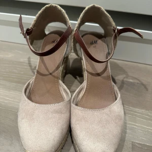 Beige kilklackar från H&M med brunt spänne - Säljer ett par beige kilklackar från H&M med öppen häl och brunt ankelband. Skorna har rund tå och ovandel i mockaliknande material samt flätad sula i rep. Perfekta för vår och sommar!