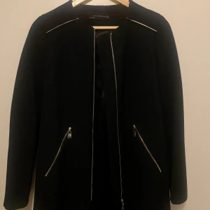 Svart kappa från Zara - Stilren svart kappa från Zara med långa ärmar och dragkedjor vid fickorna. Klassisk rak modell utan krage, perfekt för en clean look. Strl XS