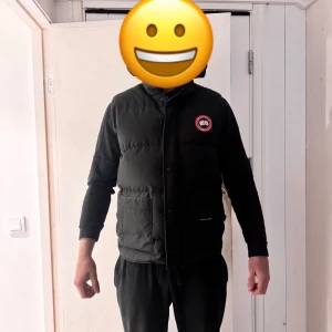 Svart dunväst från Canada Goose - Säljer en svart dunväst från Canada Goose med klassisk logotyp på bröstet. Västen har hög krage, knappar framtill och två stora fickor. Perfekt för kyliga dagar när du vill hålla dig varm och stilsäker. Nypris ligger ligger runt 5500-6000kr. På bilden är de en kille som är 188cm lång o väger runt 90kg
