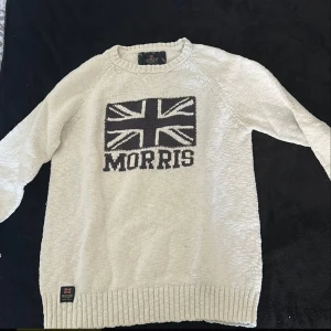 Beige stickad tröja från Morris - Säljer en beige stickad tröja från Morris med Union Jack-flagga och MORRIS-text på bröstet. Tröjan har rund halsringning och långa ärmar med ribbade muddar och nederkant. Perfekt för dig som gillar klassisk stil med brittisk touch.