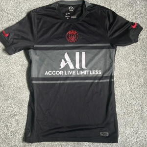 Svart PSG 21/22 fotbollströja från Nike - Säljer en svart PSG fotbollströja från Nike med röda detaljer och klubbmärke på bröstet. Tröjan har korta ärmar, Dri-FIT material och tryckt sponsorlogga. Tröjan är i princip perfekt skick förutom ett litet fel (bild 4). Köparen står för frakt. Medela mig om du har några frågor!