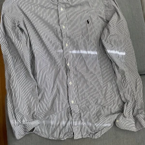 Randig skjorta från Ralph Lauren - Säljer en klassisk vit och svart randig skjorta från Ralph Lauren i storlek XL. Skjortan har långa ärmar, knappar framtill och den ikoniska broderade loggan på bröstet. Perfekt för dig som gillar stilrena plagg med tidlös design.