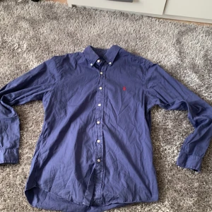 Blå skjorta från Ralph Lauren - Säljer en klassisk blå skjorta från Ralph Lauren i slim fit-modell. Skjortan har långa ärmar, knappar framtill och en röd broderad logga på bröstet. Tillverkad i tunn bomull för en lätt och bekväm känsla.