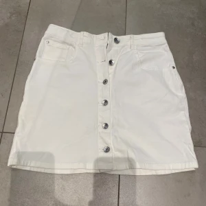 Vit jeanskjol med knappar framtill - Säljer en vit jeanskjol från Ü med snygga silverfärgade knappar längs hela framsidan. Kjolen har två fickor fram och två bak, samt hällor för bälte. Perfekt till sommaren och enkel att matcha med olika toppar.