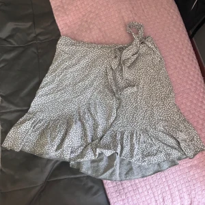 Grön omlottkjol med blommigt mönster - Supersöt grön omlottkjol, köpt i Spanien, med små vita blommor och volangkant. Kjolen har knytband men också resår där bak. Passar mig som är S men skulle också passa någon i strl M. Skriv för fler bilder och priser går att diskuteras. (Ser grå ut på bilden är typ sage grön irl) 