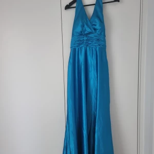Blå långklänning festklänning  - Snygg blå långklänning i glansigt material med v-ringning och omlott upptill. Haltterneck modell med dragkedja bak. Klänningen har breda axelband och rynkade detaljer vid midjan. Perfekt för bal eller festliga tillfällen. Ej använd finns dock liten fläck/färgskiftning på ena axelbanden 
