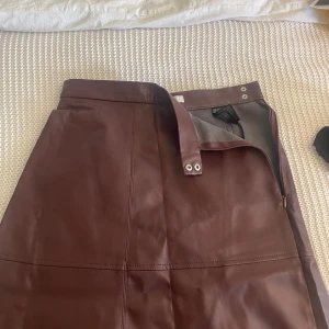 Vinröd skinnkjol från H&M - Säljer en lång vinröd skinnkjol från H&M i storlek 38. Kjolen har en rak passform, synliga sömmar och stängs med dragkedja och tryckknappar i midjan. Känns som en 36/38