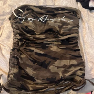 Camo tubklänning med strassdetalj - Säljer en tight tubklänning i camouflage-mönster med snörning i sidorna och strassdetaljer framtill. Klänningen har en kort passform och är perfekt för dig som vill sticka ut.