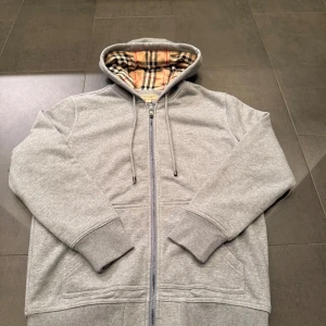 Grå hoodie med dragkedja  - Säljer en grå hoodie med klassiskt rutigt foder i huvan. Tröjan har dragkedja framtill, två fickor och ribbade muddar. Perfekt för dig som gillar stilrena detaljer och exklusiva märken.