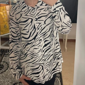 Vit och svart zebramönstrad skjorta - Säljer en snygg vit skjorta med svart zebramönster. Skjortan har långa ärmar och knäppning framtill. Perfekt för dig som vill sticka ut lite extra med din stil!