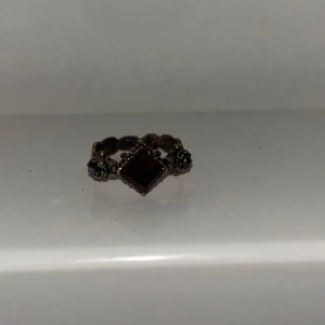 Ring med röd sten och antik design - Unik ring med en fyrkantig röd sten i mitten och detaljerade blommönster på sidorna. Ringen har en antik känsla med mörkare metall och små dekorativa detaljer runt om stenen.