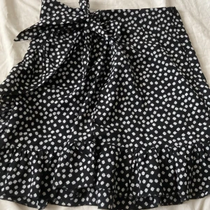 Svart blommig omlottkjol med volang från Gomorn - Supersöt svart omlottkjol från Gomorn med vita blommor, volangkant och stor rosett i midjan. Kjolen har en kort längd och är perfekt för dig som gillar en feminin och lekfull stil.