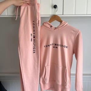 Ljusrosa hoodie från Tommy Hilfiger - Säljer en ljusrosa hoodie från Tommy Hilfiger med svart logotyp och text 'EST. 1985' på bröstet. Tröjan har huva med dragsko och en stor magficka. Perfekt för en avslappnad stil. Hela setet för 150