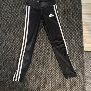 Svarta träningstights från Adidas med vita ränder - Svarta Adidas tights med klassiska vita ränder längs sidorna och Adidas-logga på låret. Modellen är tight och tillverkad i ett stretchigt funktionsmaterial (Climalite) som andas. Perfekta för träning eller sportiga outfits. Passar för 7-8 åringar! För ENDAST 75kr så får du de bästa Adidas byxorna! Pris kan diskuteras. Inga defekter. Överraska din dotter eller en nära och kära med dem här byxorna!