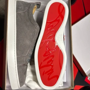 Gråa loubs - Säljer ett par gråa sneakers från Christian Louboutin med ovandel i mocka och klassisk röd sula. Skorna har snörning och vit platt sula. Perfekta för dig som vill ha en stilren och lyxig look.