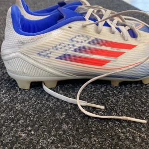 Adidas fotbollsskor med röda och blå detaljer - Säljer ett par Adidas fotbollsskor i vitt med röda och blå detaljer. Skorna har snörning och är designade för att ge bra grepp på planen med sina röda dobbar. Insidan är blå och de har en sportig look.