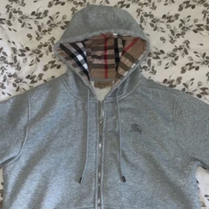 Burberry zip  - Snygg grå hoodie från Burberry med klassiskt rutigt foder i huvan. Tröjan har dragkedja framtill, justerbar huva med snören och är tillverkad i mjukt bomullsmaterial. Perfekt för en avslappnad och stilren look.
