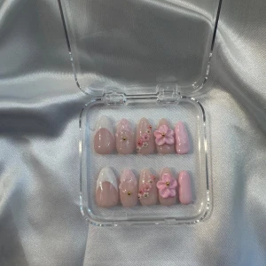 Rosa press-ons  med 3D-blommor och pärldekor - Säljer ett set med rosa press-ons i olika nyanser och former. Naglarna har detaljer som vita tippar, 3D-blommor, små pärlor och guldiga dekorationer. Perfekt för dig som vill ha en söt och unik nageldesign.