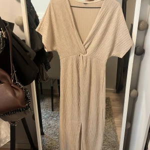 Beige ribbad omlottklänning - Säljer en beige ribbad klänning med omlott-design och v-ringning. Klänningen har korta ärmar och en knytning i midjan som ger en snygg siluett. Perfekt för dig som gillar en stilren och avslappnad look. Från nakd 