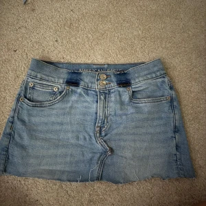 Ljusblå jeanskjol från American Eagle - Säljer en ljusblå jeanskjol från American Eagle med rå kant nertill och slitna detaljer på bakfickorna. Kjolen har klassisk femficksdesign och dubbla knappar framtill. Perfekt till sommaren!